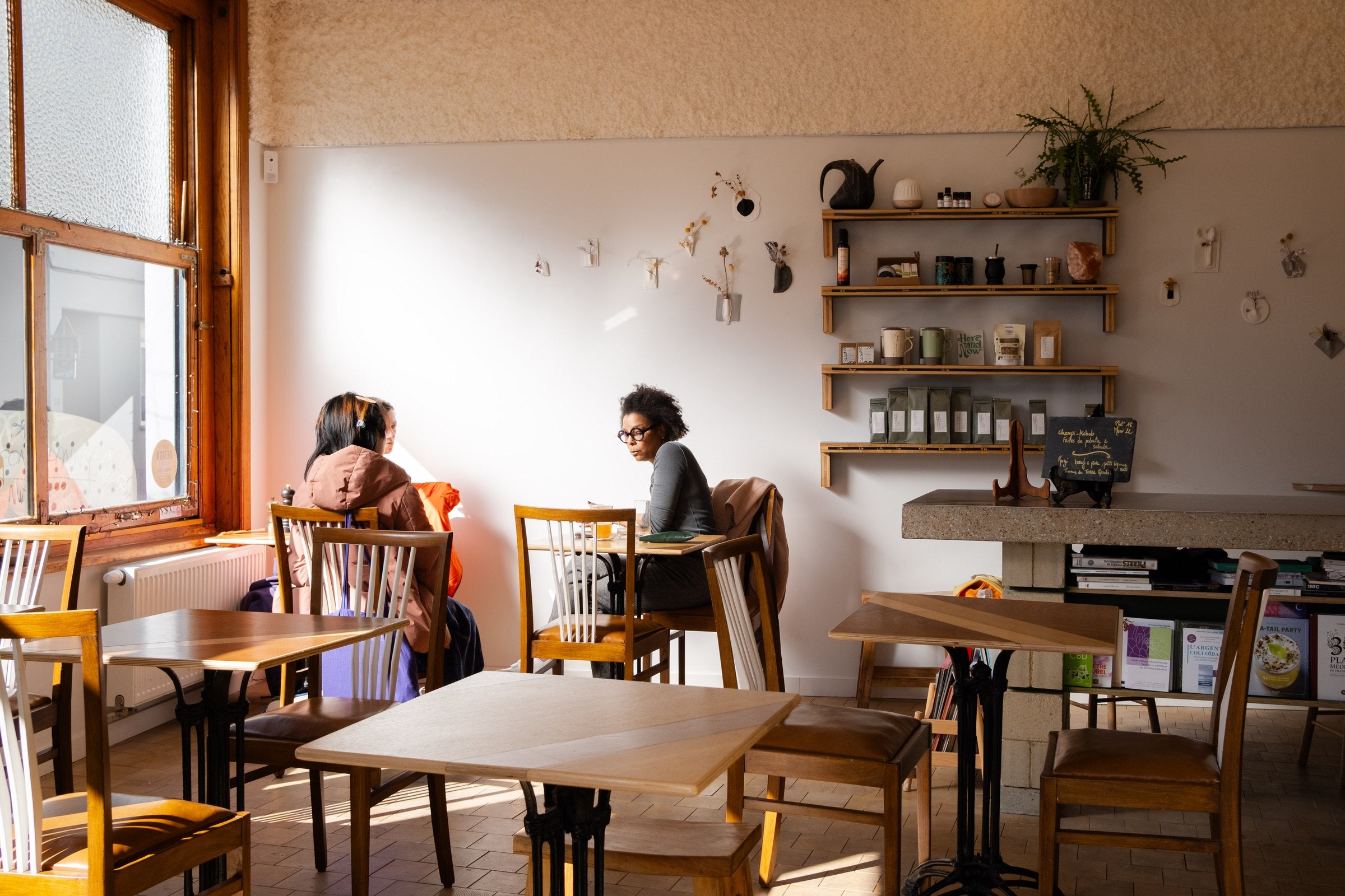 La salle de Place aux Plantes à Ixelles, baignée de lumière, avec ses tables en bois et les étagères du comptoir d'herboristerie